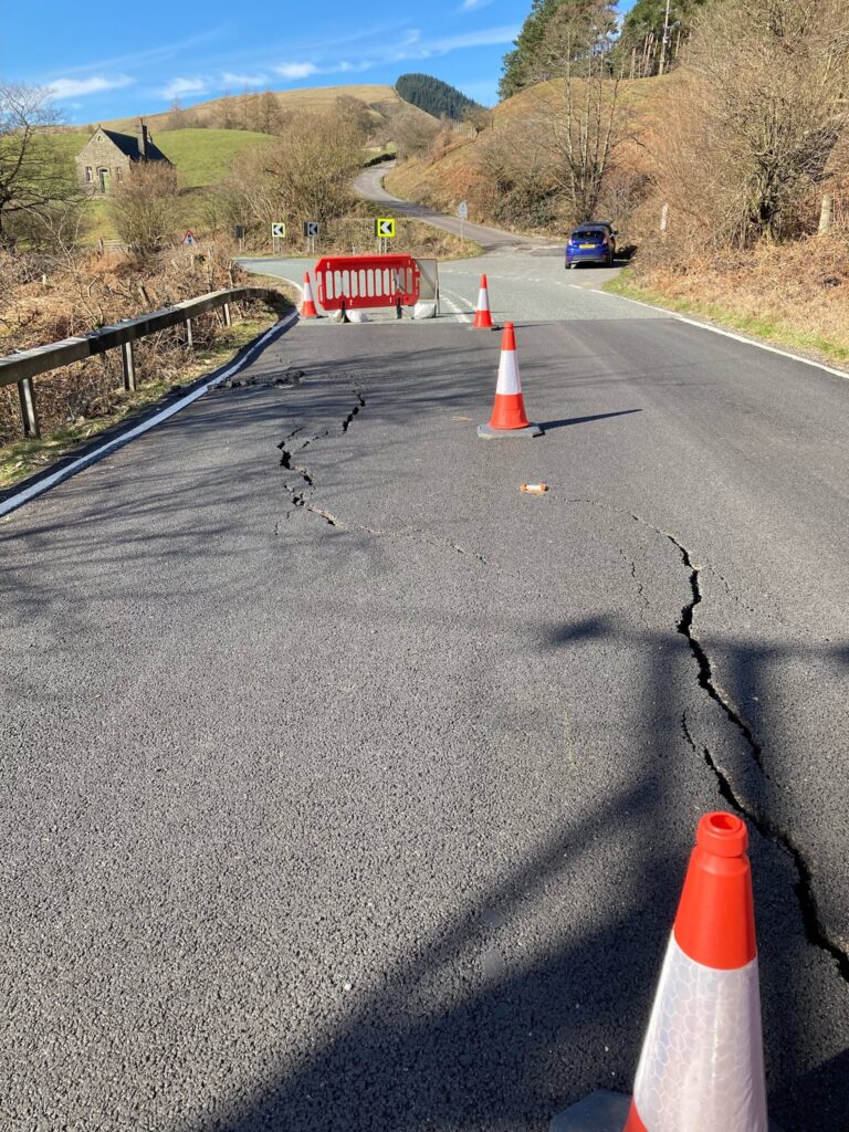 Snake road, on a bend with traffic cones, and one side of road has big cracks plus is sunken 